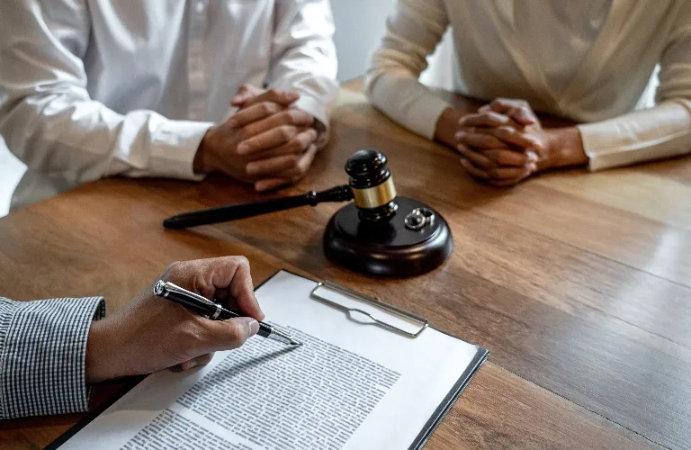 Two individuals with folded hands sit at a wooden table with a judge's gavel, while a third person reviews and marks a document on a clipboard.