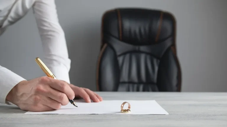 Person signing a document on a desk with a gold wedding ring placed on the paper.