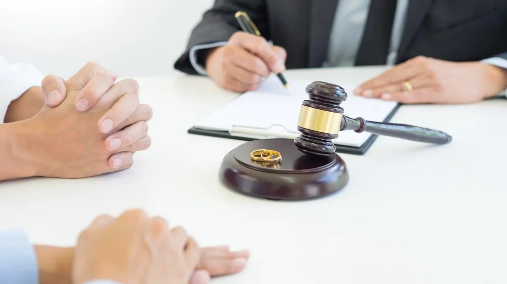 Close-up of a judge's gavel, wedding rings, and two people with a lawyer signing documents.