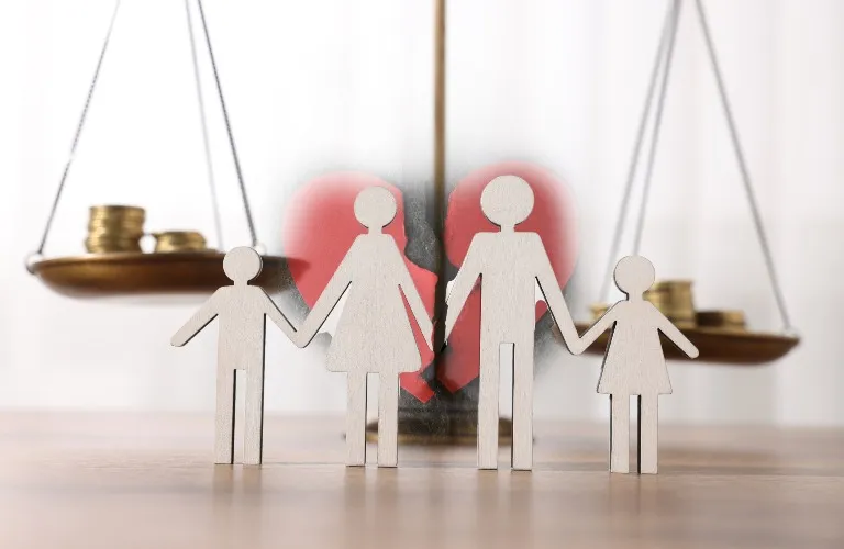 Wooden cutout figures of a family in front of a broken red heart and balanced scales with coins.