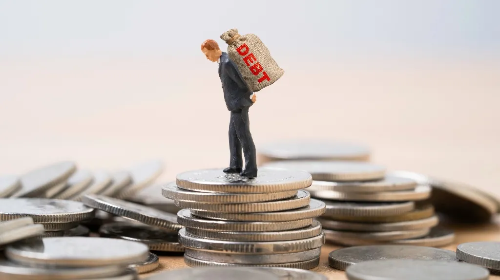 Miniature figurine of a man in a suit carrying a sack labeled "DEBT" on stacked silver coins.