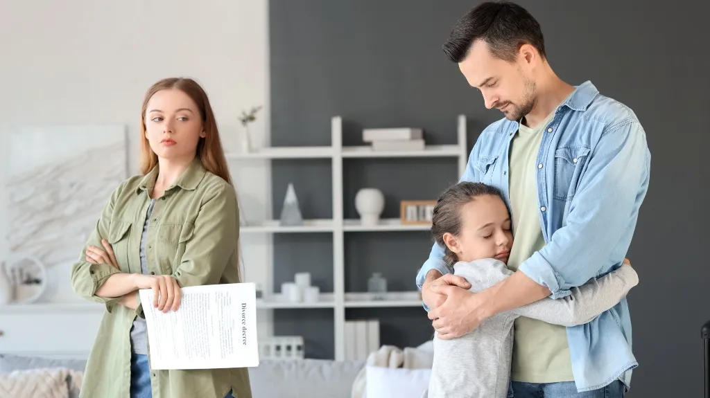 Woman holding divorce papers with arms crossed, man hugging a child in living room.