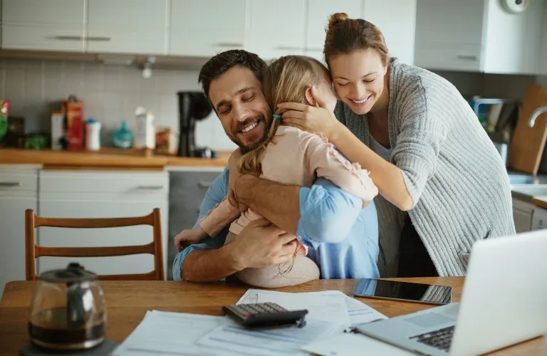 A smiling couple embraces their young child in a modern kitchen with documents and a laptop on the table.