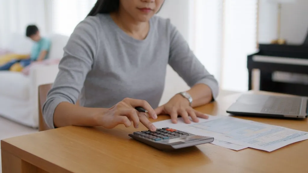 Woman in gray shirt using calculator at wooden table with documents and laptop.