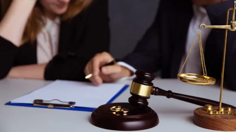 Close-up of a judge's gavel, wedding rings, and scales of justice on a desk with two people blurred in background.