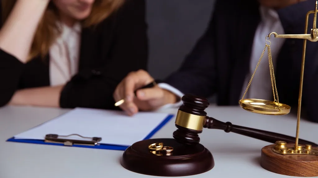 Close-up of a judge's gavel, wedding rings, and scales of justice on a desk with two people blurred in background.