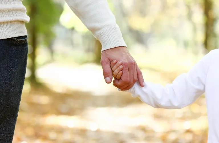 Adult and child holding hands outdoors, both wearing white long-sleeve tops.